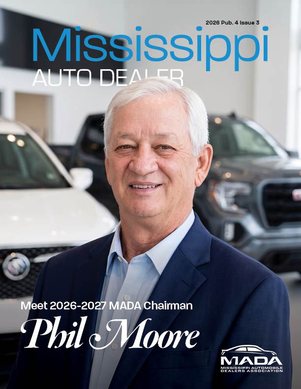 Phil Moore, white hair and light skin, Standing in his dealership showroom in a light blue button up shirt ad a dark blue suit jacket.